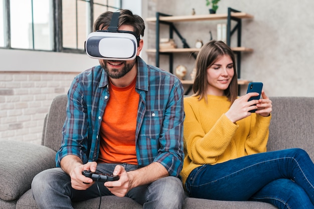Молодой человек в очках виртуальной реальности играет в видеоигру с женой с помощью мобильного телефона