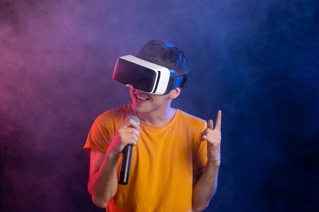 Young man wearing virtual reality headset and holding mic dark blue surface