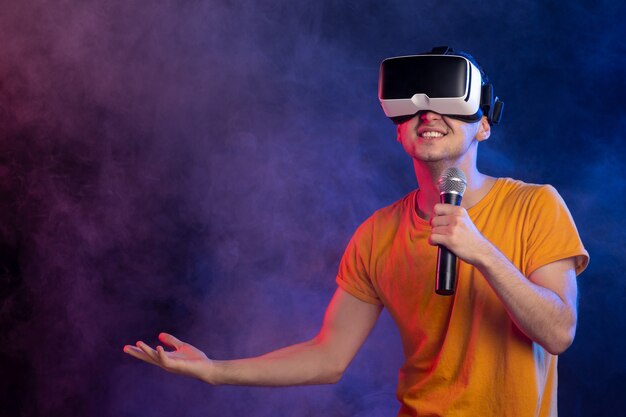 Young man wearing virtual reality headset and holding mic dark blue surface