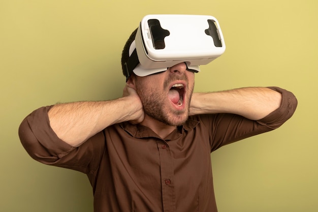 Free photo young man wearing vr headset keeping hands behind neck looking at side screaming isolated on olive green wall