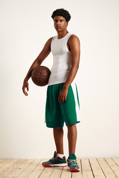 Young man wearing a white shirt, headband, green shorts and green sneakers standing with an old leather basketball under his arm