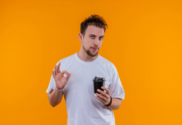 молодой человек в белой футболке держит чашку кофе, показывая жест окей на изолированной оранжевой стене