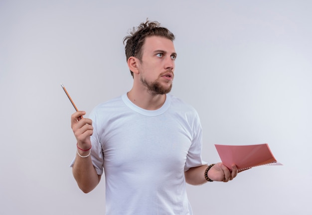  young man wearing white t-shirt holding pencil and notebook on isolated white wall