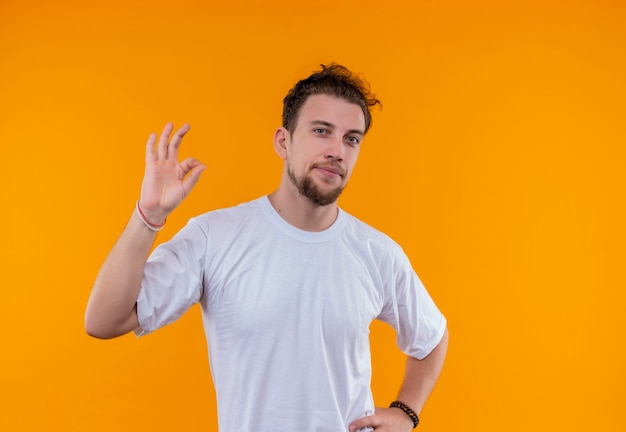 孤立したオレンジ色の壁に大丈夫なジェスチャーを示す白いTシャツを着た若い男