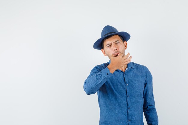 Young man whistling with fingers in blue shirt, hat, front view.