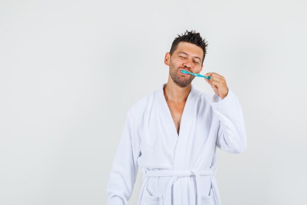 Young man in white bathrobe brushing teeth with closed eyes , front view.