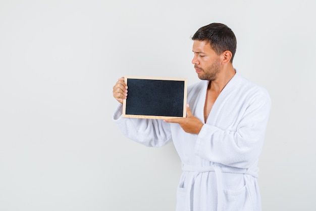 Young man in white bathrobe holding blackboard and looking pensive , front view.