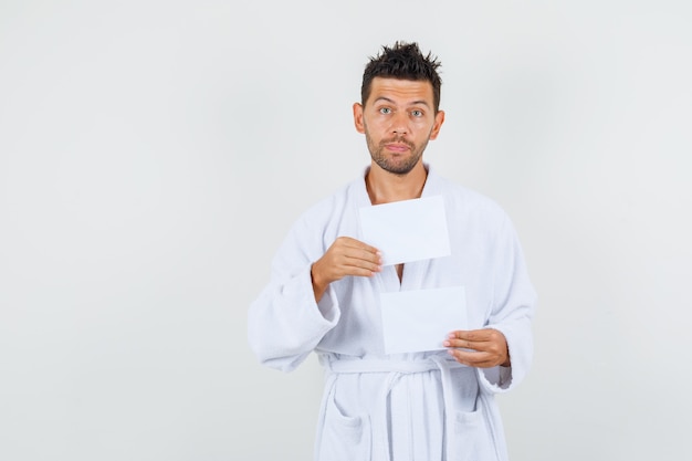 Young man in white bathrobe holding blank paper sheets , front view.