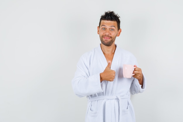 Young man in white bathrobe holding cup of tea with thumb up and looking cheerful , front view.