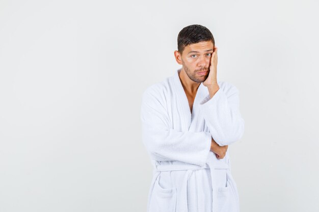 Young man in white bathrobe holding palm on cheek and looking pensive , front view.
