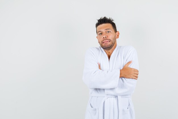 Young man in white bathrobe hugging himself and feeling cold , front view.