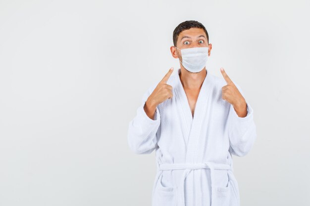 Young man in white bathrobe pointing at mask and looking careful , front view.