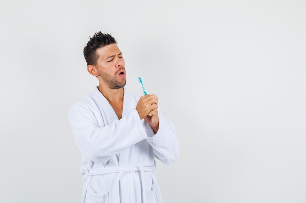 Young man in white bathrobe singing into toothbrush like microphone and looking funny , front view.