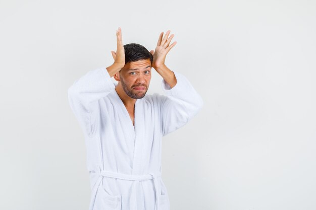 Young man in white bathrobe touching head with palms and looking sad , front view.