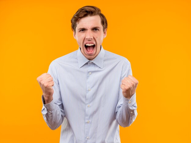 Молодой человек в белой рубашке, сжимая кулаки, разочарованный и сердитый с агрессивным выражением лица стоит над оранжевой стеной