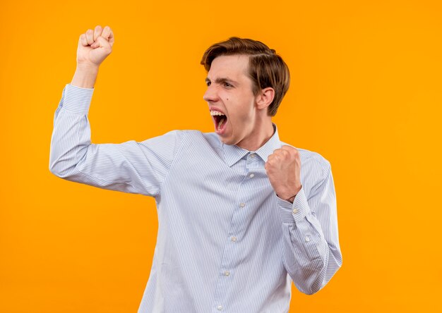 Young man in white shirt clenching fists happy and excited shouting standing over orange wall