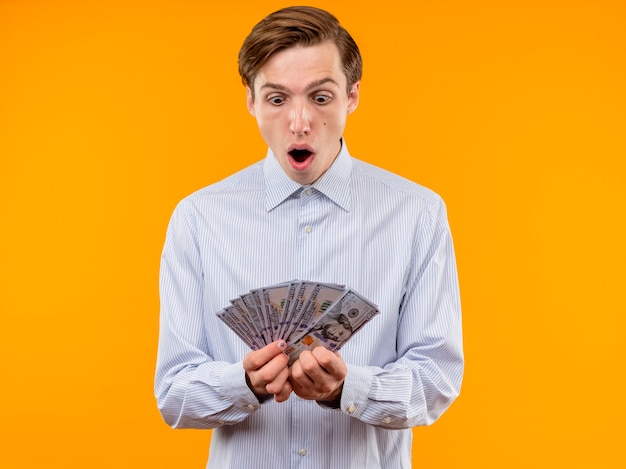 Young man in white shirt holding cash looking amazed and surprised standing over orange wall