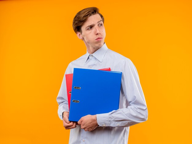 Young man in white shirt holding folders looking aside with serious face standing over orange background