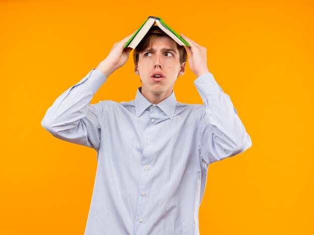 Young man in white shirt holding open book over his head looking aside scared standing over orange wall