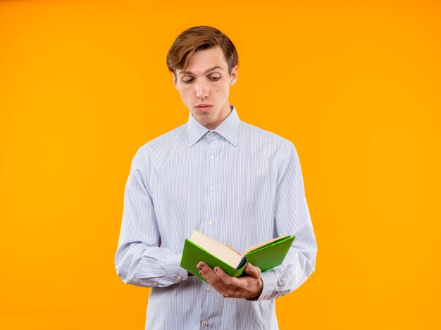 Young man in white shirt holding open book lookign at it reading with skeptic expression standing over orange background