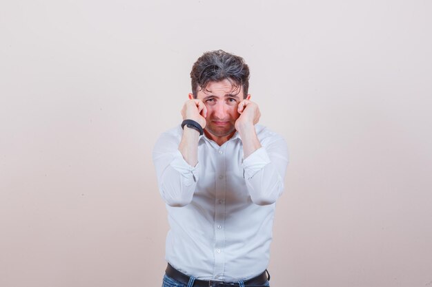 Young man in white shirt, jeans plugging ears with fingers and looking annoyed