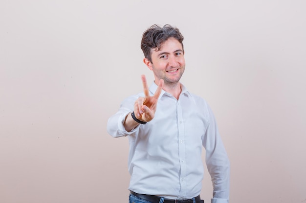 Young man in white shirt, jeans showing two fingers ow v-sign and looking cheery