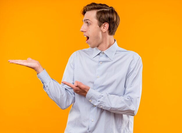 Young man in white shirt looking aside surprised and happy presenting something with arm of his hand pointing with index finger at something standing over orange background