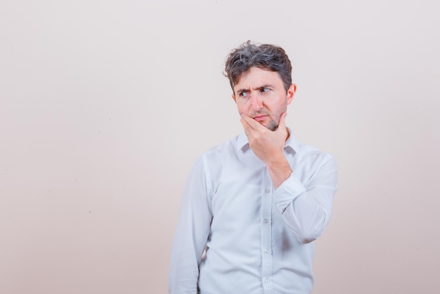 Young man in white shirt looking aside with hand on chin and looking thoughtful