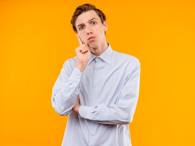 Young man in white shirt looking at camera with pensive expression expression on face standing over orange background