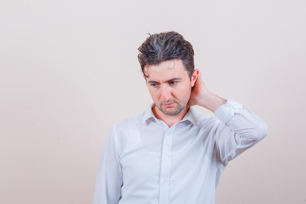 Young man in white shirt looking down with hand on neck and looking pensive