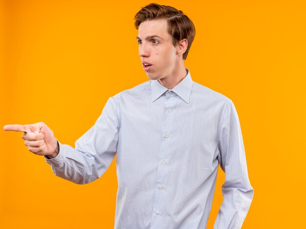 Молодой человек в белой рубашке указывает указательным пальцем на что-то растерянное, стоя у оранжевой стены