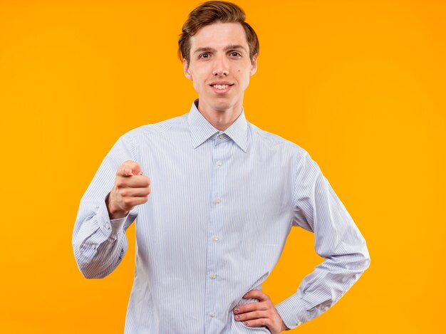 Молодой человек в белой рубашке, указывая указательным пальцем, уверенно улыбаясь, стоит над оранжевой стеной