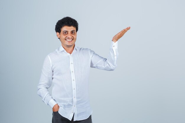 Молодой человек в белой рубашке показывает что-то выше или приветствует и выглядит веселым, вид спереди.