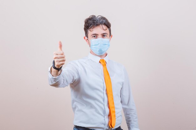 Young man in white shirt, tie, mask showing thumb up and looking pleased