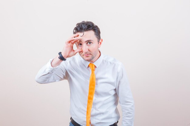 Young man in white shirt, tie opening eye with fingers and looking tired