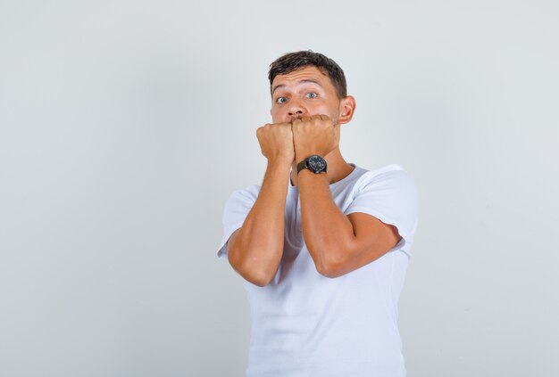 Young man in white t-shirt biting fists and looking nervous, front view.