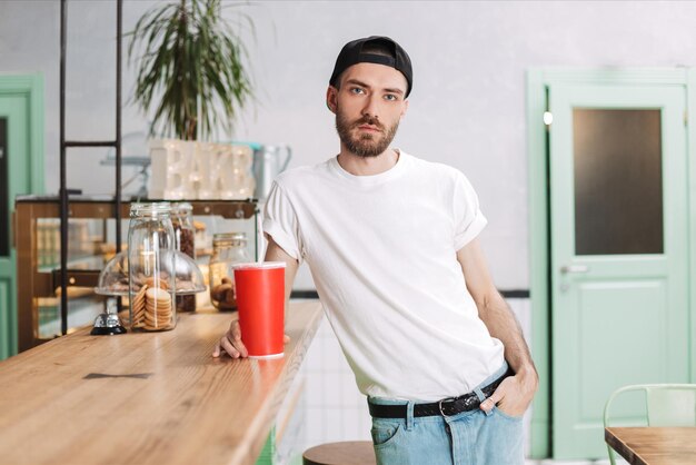 Young man in white t shirt and black cap standing at the bar counter with soda water and dreamily looking in camera in cafe