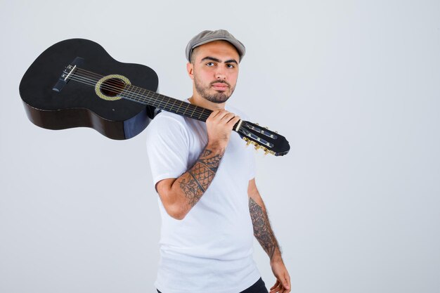 Young man in white t-shirt, black pants, grey cap holding guitar and looking serious