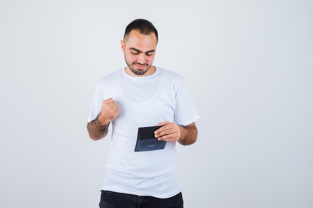 Young man in white t-shirt and black pants holding calculator and clenching fist and looking serious