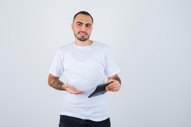 Young man in white t-shirt and black pants holding calculator and pointing to it and looking happy