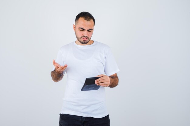 Young man in white t-shirt and black pants holding calculator and stretching hand in questioning manner and looking perplexed