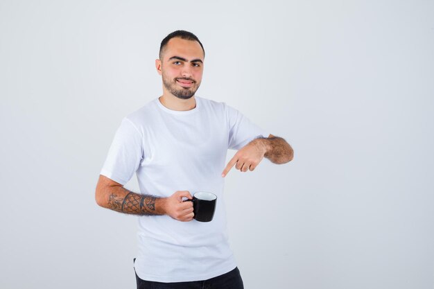 Young man in white t-shirt and black pants holding cup of tea and pointing to it and looking happy