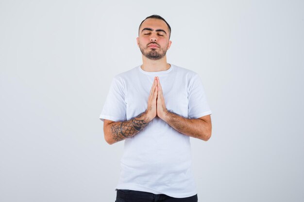 Young man in white t-shirt and black pants showing namaste gesture and looking serious