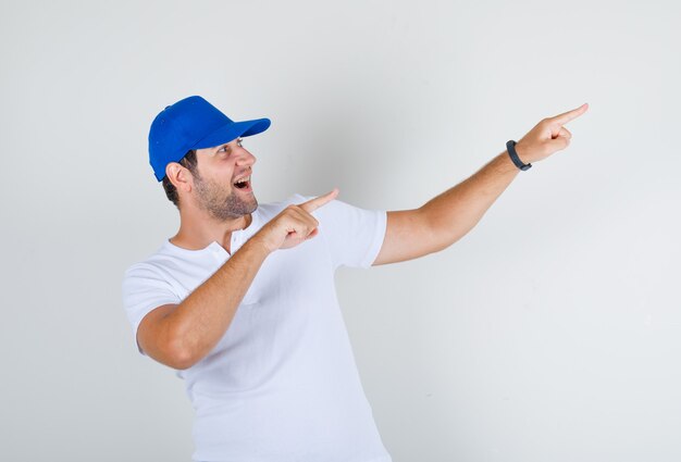 Young man in white t-shirt, blue cap pointing fingers away and looking happy