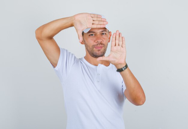 Young man in white t-shirt, cap doing frame gesture and smiling, front view.