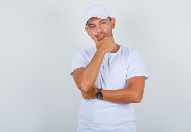 Young man in white t-shirt, cap holding hand on chin and looking confident, front view.
