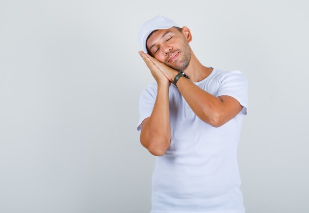 Young man in white t-shirt, cap leaning on palms as pillow and looking relaxed, front view.