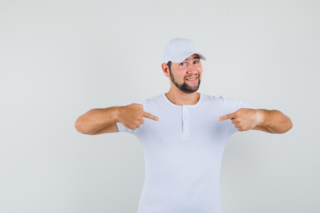 Young man in white t-shirt,cap pointing at himself and looking jolly , front view.