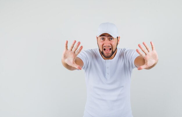 Young man in white t-shirt,cap raising hands with preventive manner and looking frightened , front view.