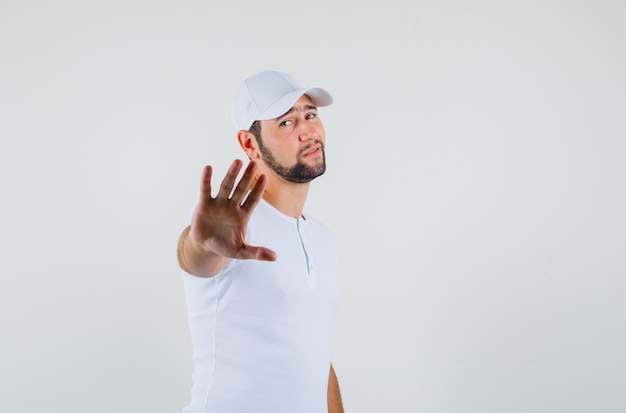 Young man in white t-shirt,cap showing stop gesture and looking sorrowful , front view. space for text